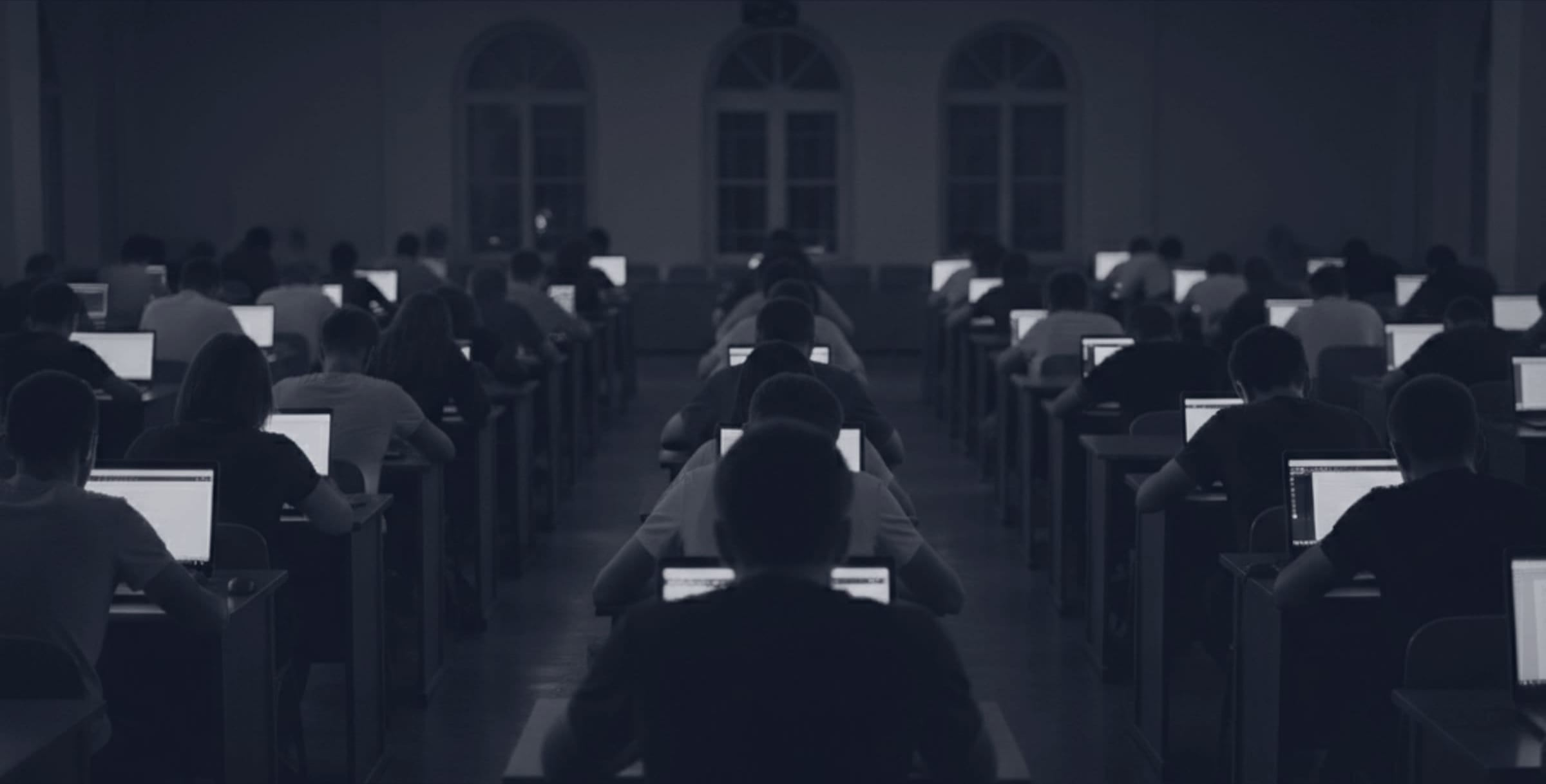 Un aula universitaria durante un examen: filas de estudiantes de espaldas con sus portátiles encendidos, en penumbra.